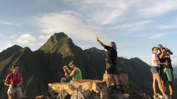Tourists stand on a mountaintop in Laos taking photos and selfies