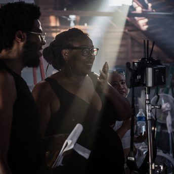 Production still of Angela Tucker looking into a camera monitor with her hand over her mouth and smiling. She is wearing glasses and has her hair in a bun. Standing next to her is a man wearing glasses and holding a notebook.