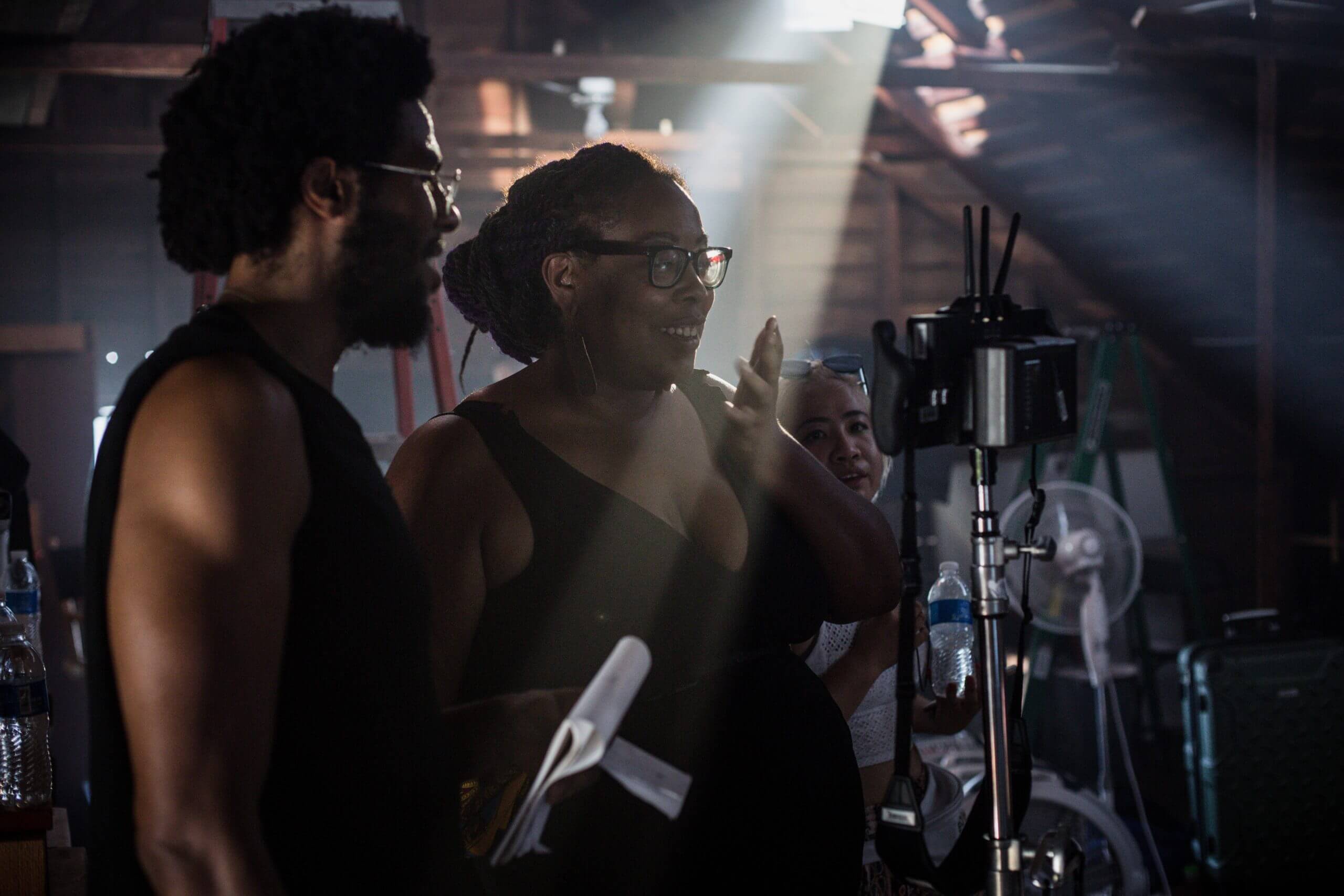Production still of Angela Tucker looking into a camera monitor with her hand over her mouth and smiling. She is wearing glasses and has her hair in a bun. Standing next to her is a man wearing glasses and holding a notebook.