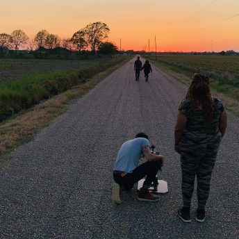 Production still of Angela Tucker standing on a road with her back to the camera. She is wearing a camouflage jumpsuit and is next to a man bending down with a camera. In the background, a couple walks off toward a setting sun.