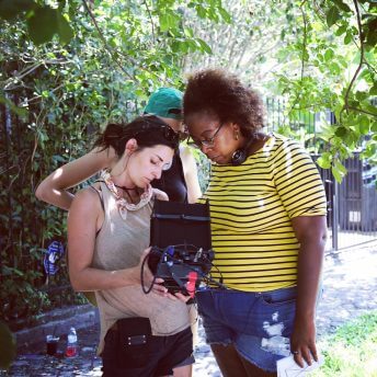 Production still of Angela Tucker in a yellow and blue striped shirt. She is standing and looking into a camera held by a woman with brown hair holding the camera underneath a tree.