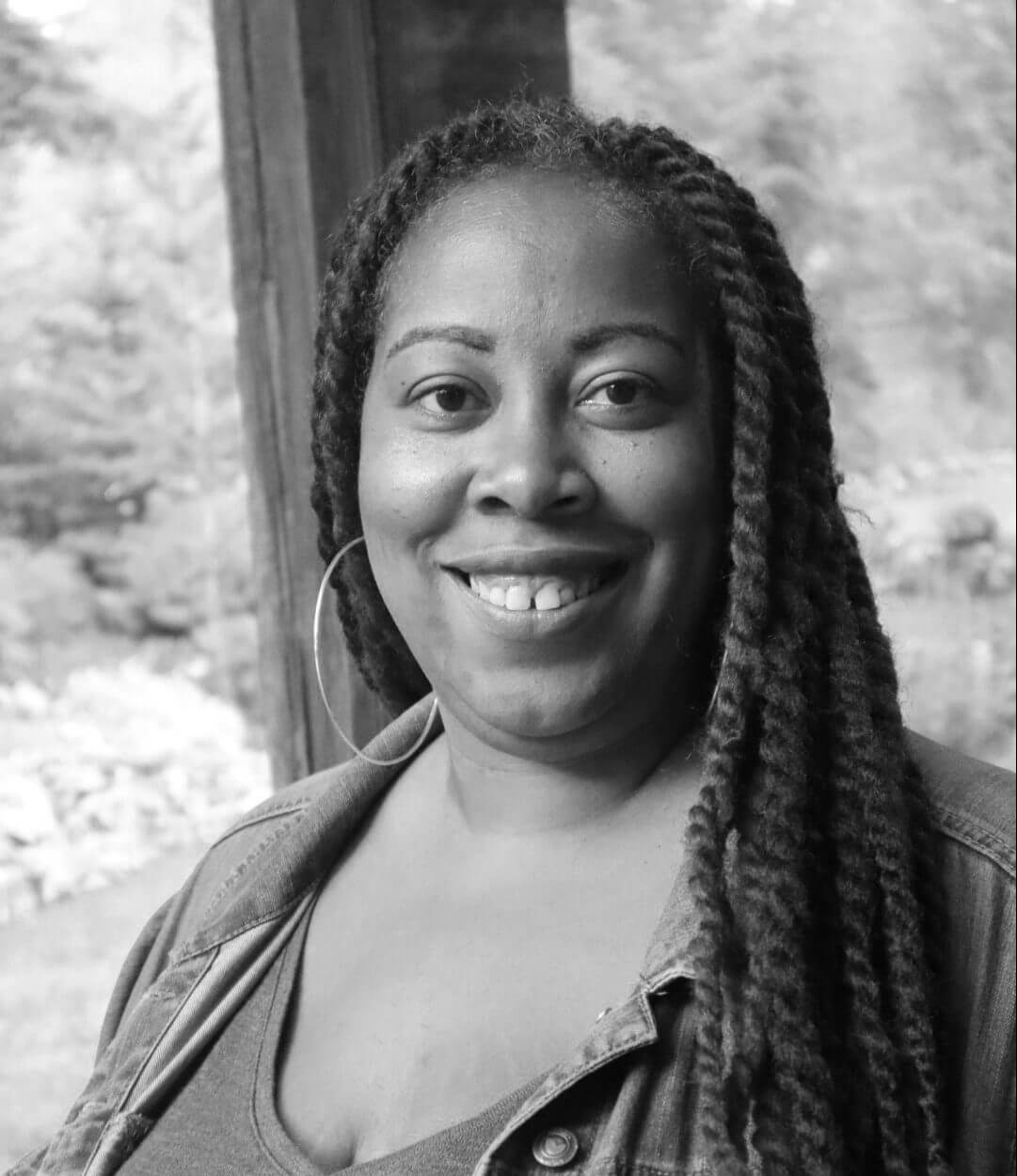 Angela Tucker looking directly at the camera and smiling. She is wearing a denim jacket, a turquoise shirt, and purple and brown crocheted dreads. Black and white portrait
