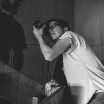 Production still of Chuck Norris vs. Communism. Ilinca Calugareanu is looking through the director’s viewfinder and is leaning over a stairwell. To her left, there is a bearded man standing on the stairs above her.