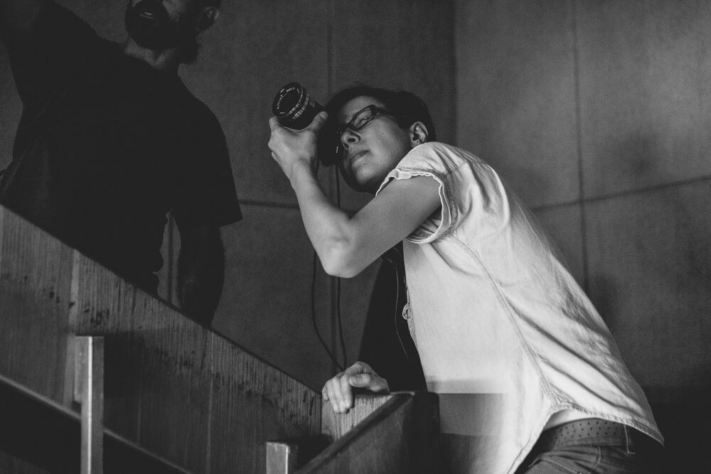 Production still of Chuck Norris vs. Communism. Ilinca Calugareanu is looking through the director’s viewfinder and is leaning over a stairwell. To her left, there is a bearded man standing on the stairs above her.