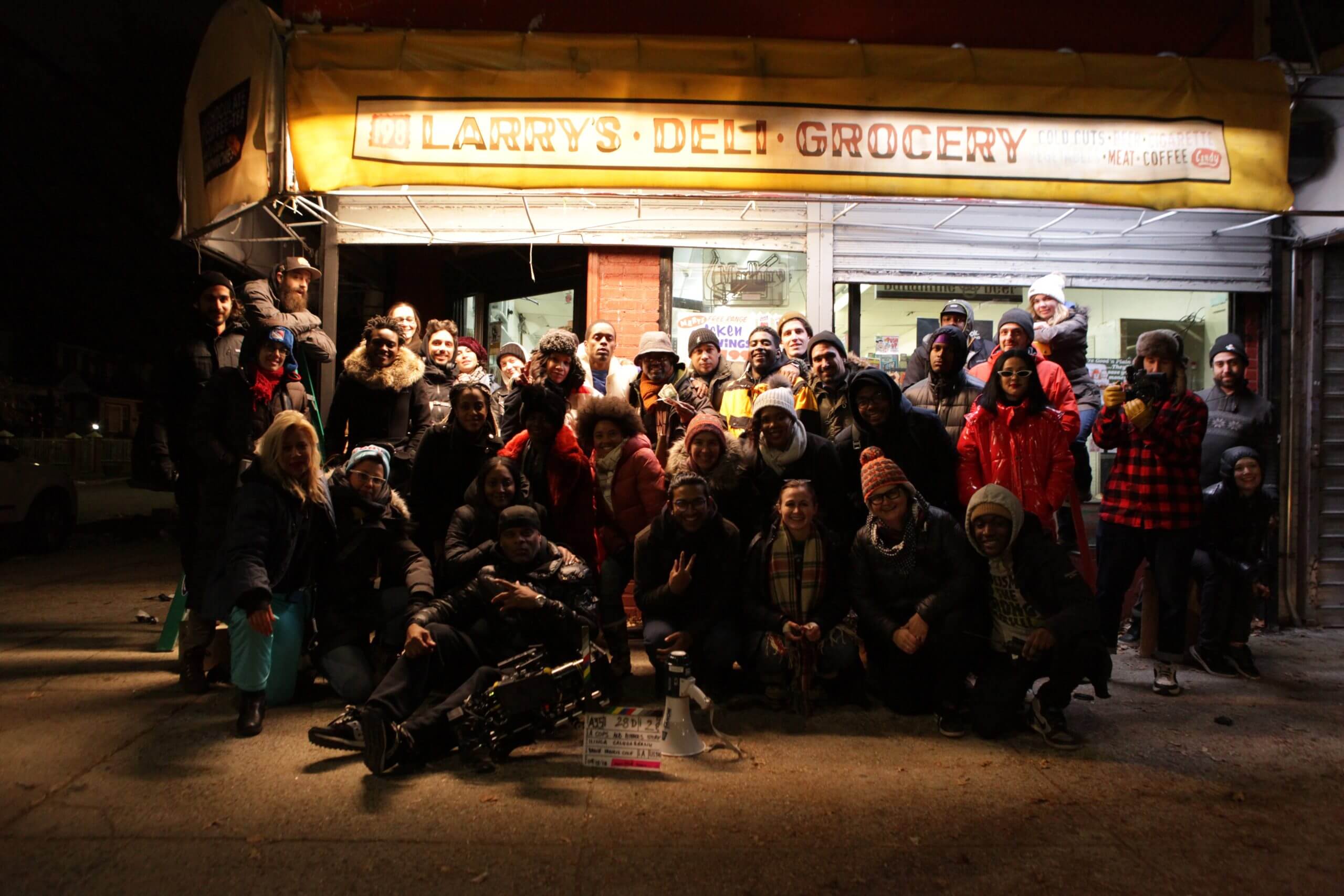Production still of A Cops and Robbers Story. Crew, cast and documentary subjects are posing for a final photo at the end of the dramatizations shoot. They are outside at night and stand underneath a bodega's lit awning that reads "Larry's Deli Grocery."