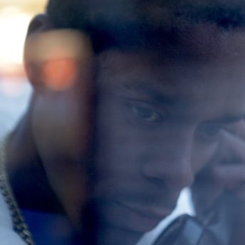Still from A Cops and Robbers Story. Closeup of teenage Corey Pegues talking on a payphone. The light from the street reflects on his face, which is focused and serious (photographer Emir Fils-Aime)