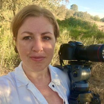 Production still of Jeanie Finlay looking directly into the camera. She has tied-back ginger hair and is wearing a white shirt. She is standing in a field in Spain with a DSLR camera on a tripod that is leaning against her shoulder.