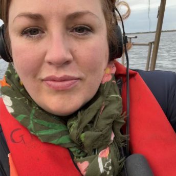 Production still of Jeanie Finlay looking directly into the camera. She is on a fishing boat on the North Sea and is operating a camera. She is wearing a green floral scarf and a life jacket.