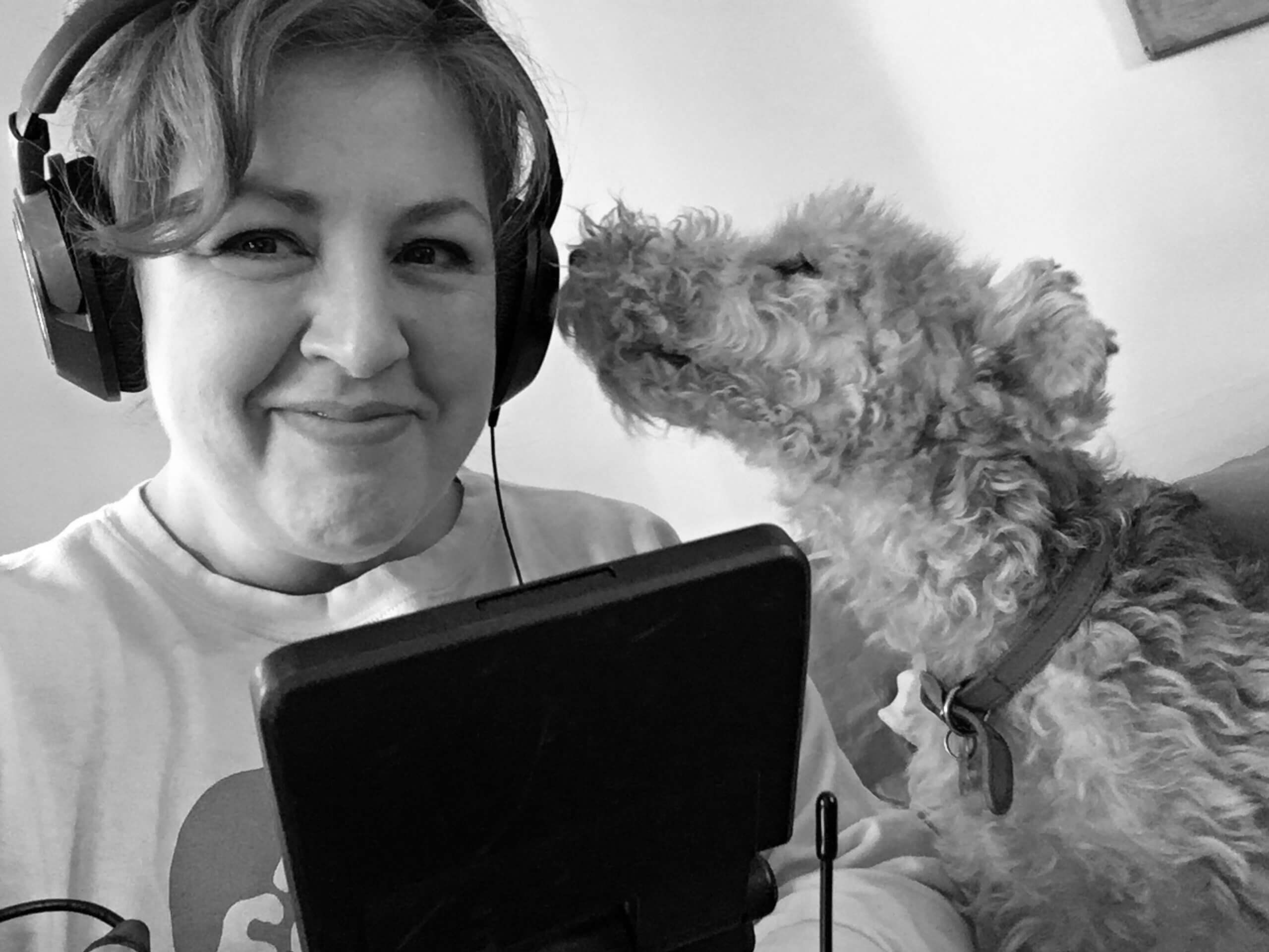 Production still of Jeanie Finlay looking directly at the camera and smiling. She is wearing headphones and holding a camera. To her right is a fox terrier dog, Dandy from the film Seahorse, who is sniffing her ear.