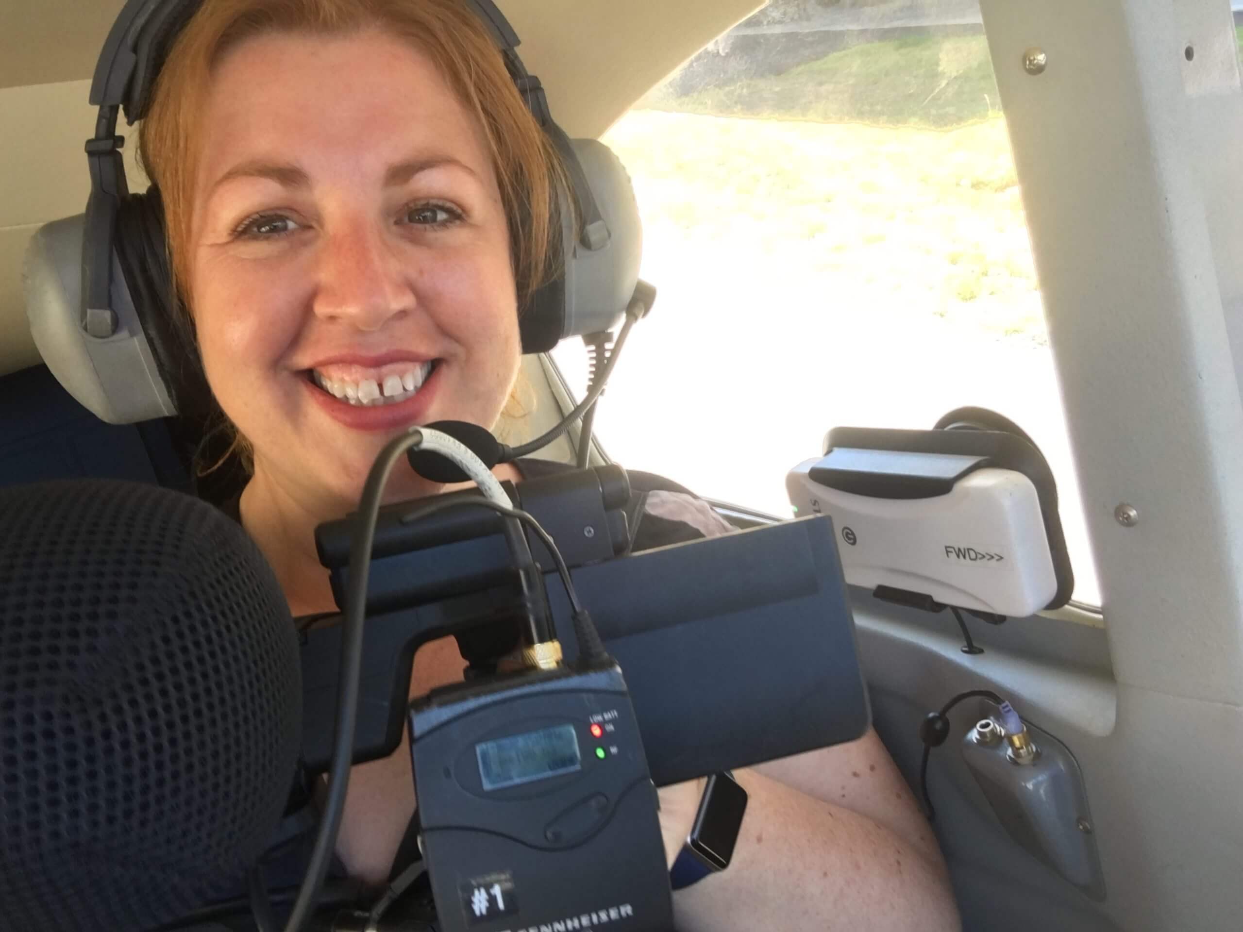 Production still of Jeanie Finlay looking directly at the camera and smiling. She is sitting in the back of a small Cessna plane, wearing headphones and holding a camera.