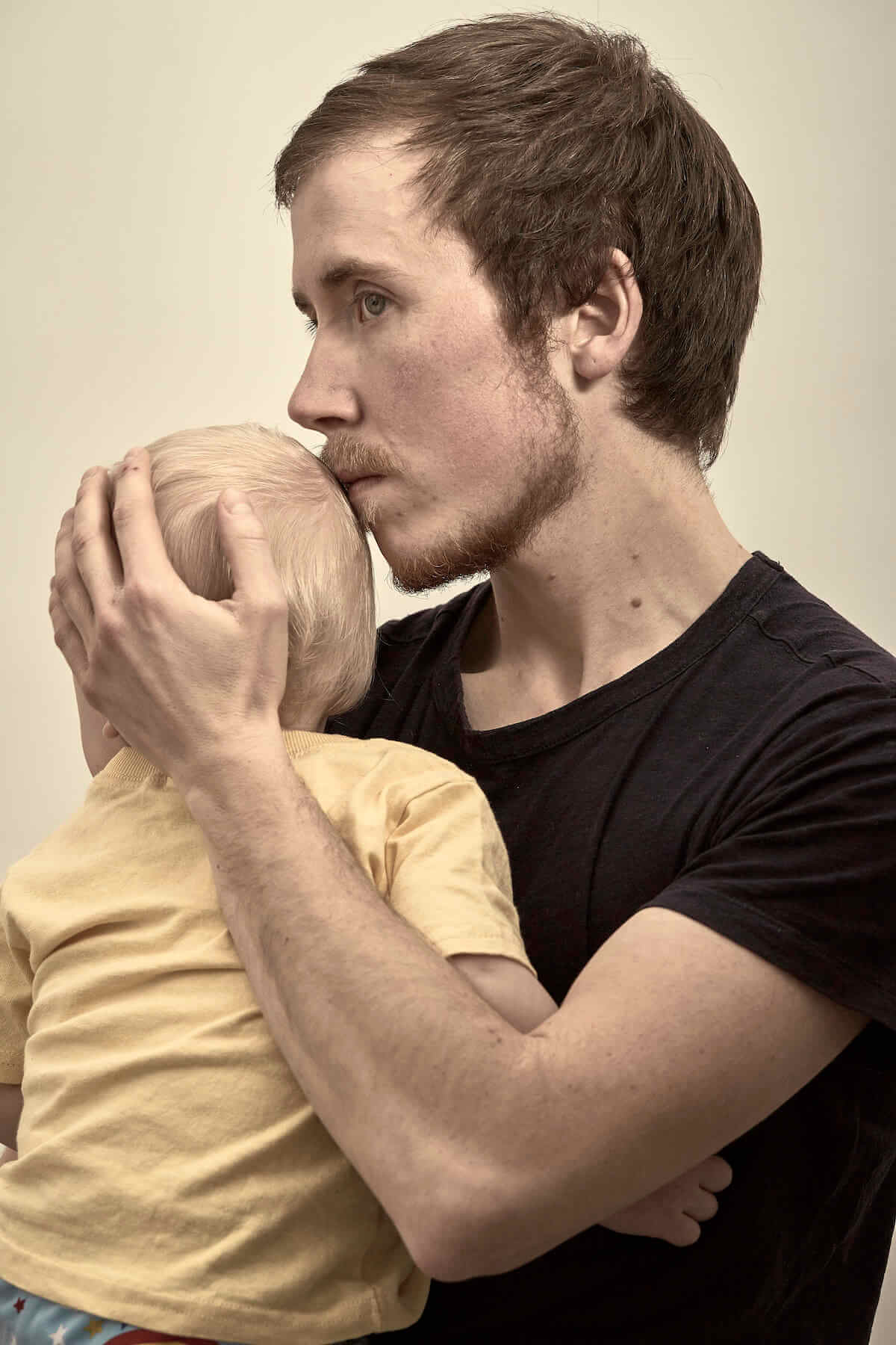 Still from Seahorse: The Dad Who Gave Birth. Freddy is standing in profile to the camera. He has short brown hair and facial hair and is cradling the son he carried and gave birth to. The child has their face turned away and you see the blonde back of their head.