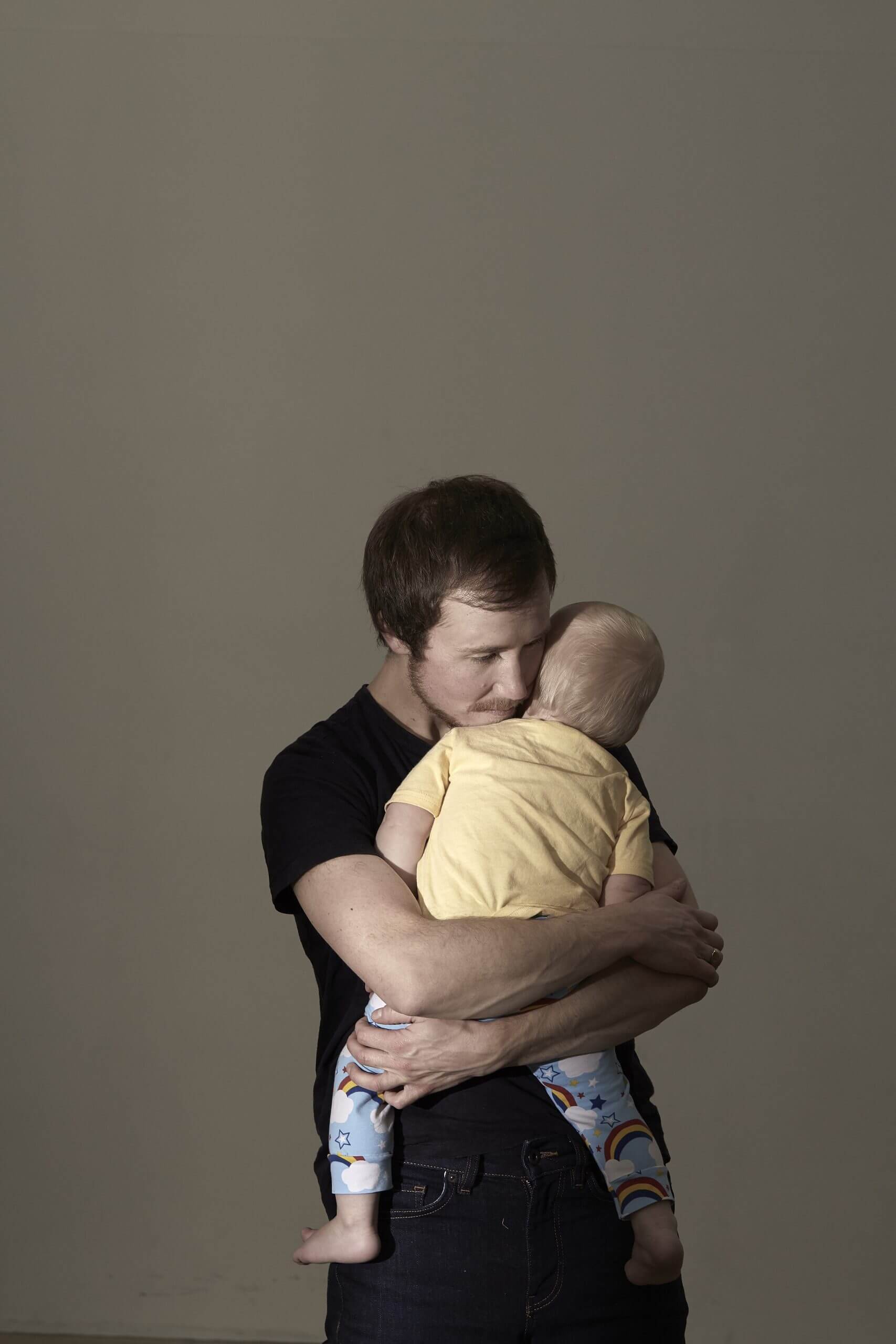 Still from Seahorse: The Dad Who Gave Birth. Freddy is standing against a green background. He is cradling the son he carried and gave birth to. The child has their face turned away and the blonde back of their head is shown.