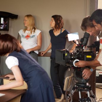 Production still of Savage. Lisa Jackson is standing amongst the crew in the background and has short brown hair. She is standing in profile and looking off at a camera monitor. Two men are at the camera, looking down and filming the shot.