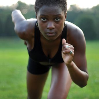 Still from Off and Running. A young woman staring directly into the camera posed to begin sprinting. She is wearing a black tank top, black shorts, and gold hoop earrings.