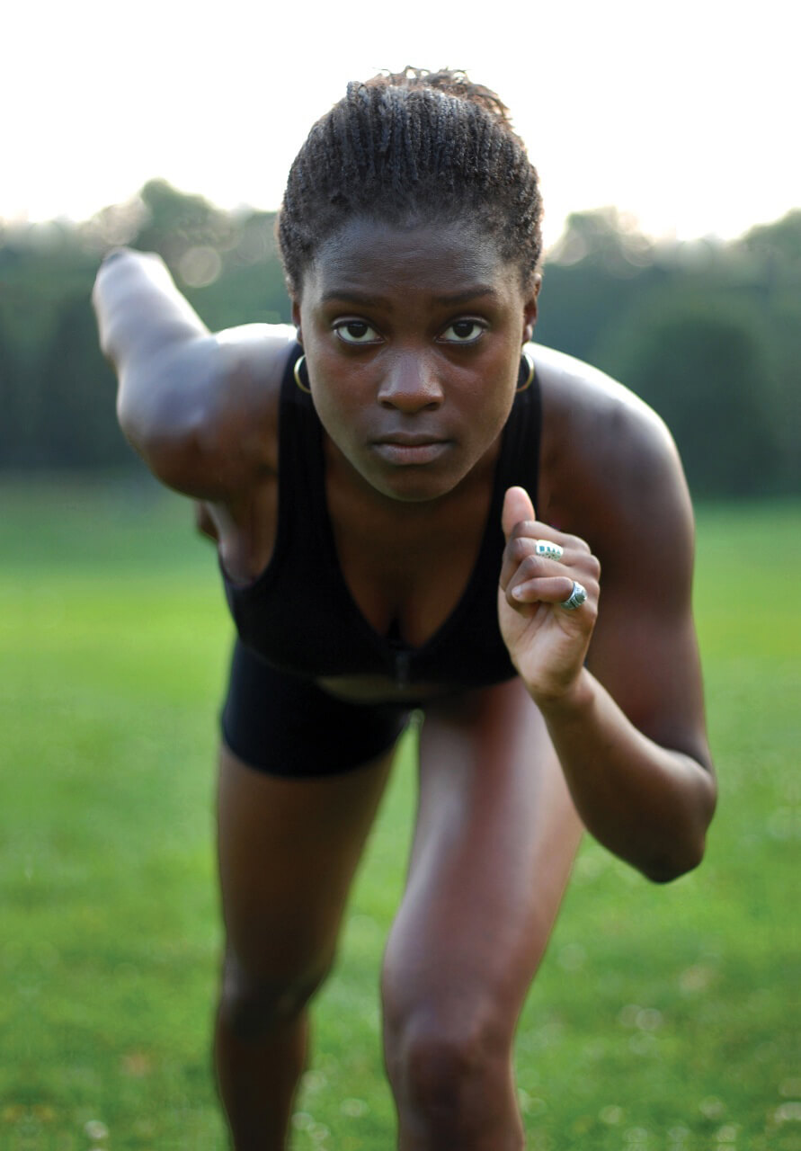 Still from Off and Running. A young woman staring directly into the camera posed to begin sprinting. She is wearing a black tank top, black shorts, and gold hoop earrings.