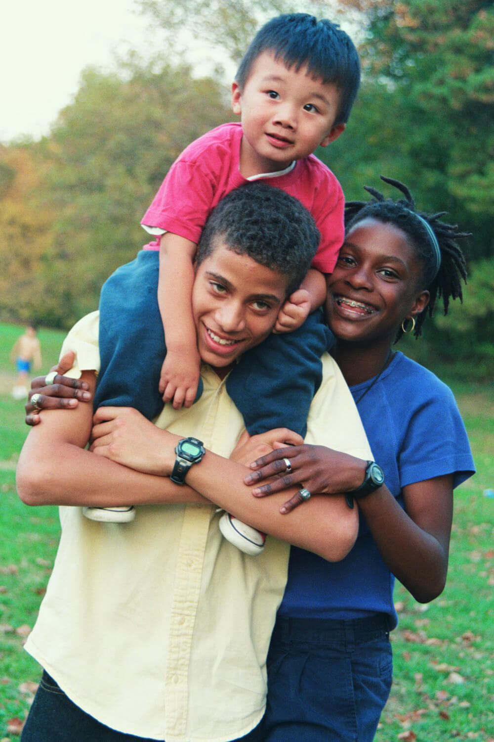 Still from Off and Running. A young girl wearing a blue t-shirt smiles into the camera with her two siblings by adoption. The youngest is wearing a red t-shirt and jeans and the oldest wearing a yellow button-up shirt. The younger sibling sits on his older brother's shoulders.
