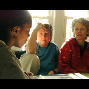 Still from Off and Running. A young woman wearing a grey sweatshirt talks with her two adoptive moms in their Brooklyn kitchen. The mom in the middle has short cropped grey hair and wears a blue t-shirt. The mom on the right wears a red knit sweater and also has short-cropped grey hair. Their expressions are somber.