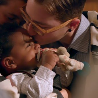 Still from The F Word. A nonbinary individual with short auburn hair and wood-framed glasses kisses their adopted Black and Filipino baby on the forehead.