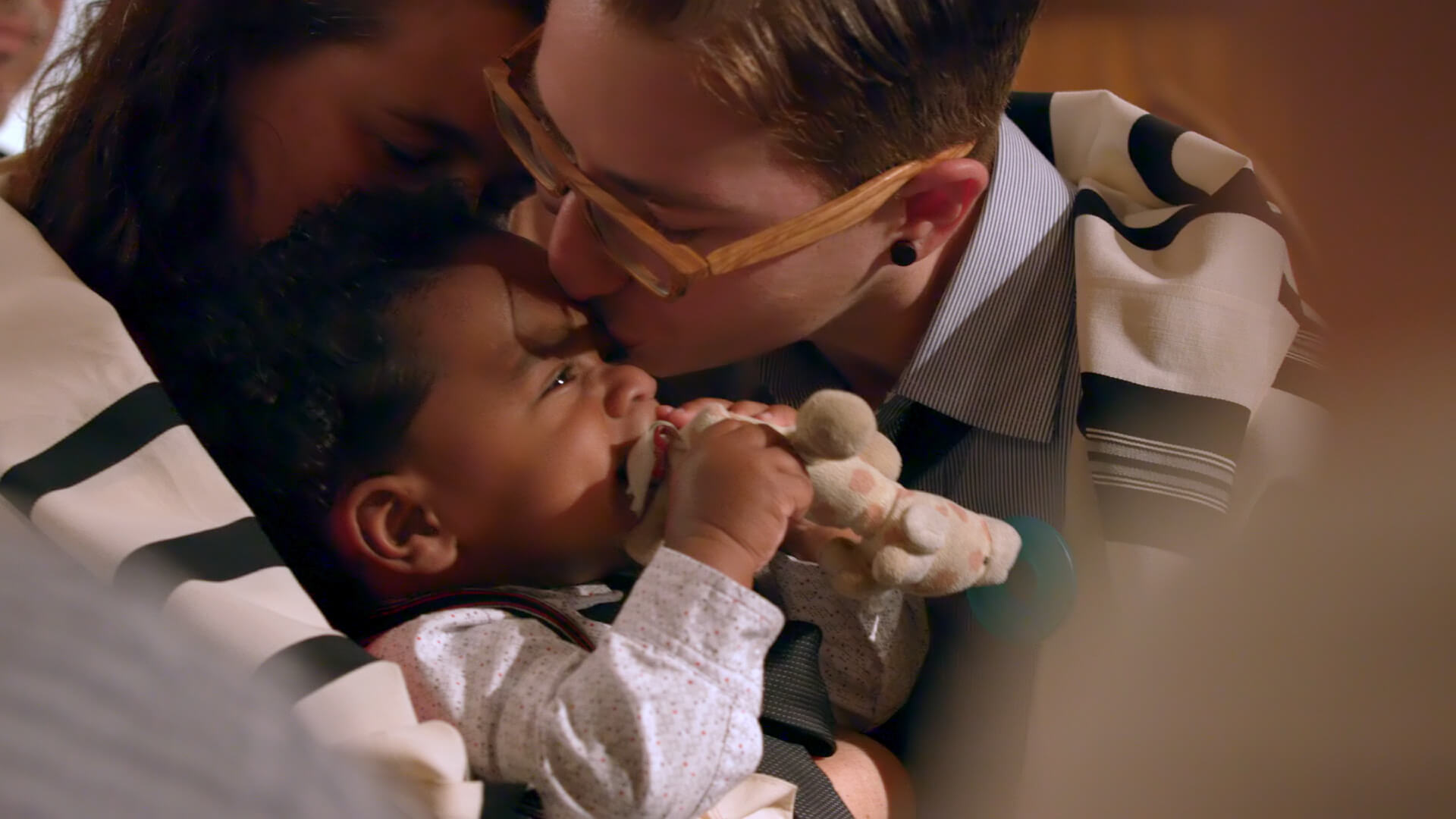 Still from The F Word. A nonbinary individual with short auburn hair and wood-framed glasses kisses their adopted Black and Filipino baby on the forehead.