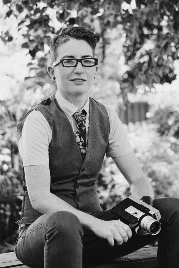 Nico Opper looking directly at the camera. They are wearing a floral tie, vest, and wood-framed earrings. They are sitting on a park bench holding a Super 8 camera. Black and white portrait.