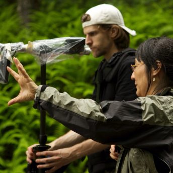 Production still of Rea Tajiri standing in a side profile to the camera. She is standing next to a man in baseball cap holding steadicam, and pointing to something in the distance.