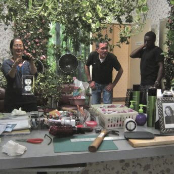 Production still of Rea Tajiri, David Wasco, and Dru Mungai looking into a long beauty shop mirror. Rea is holding a camera and photographs their reflection. Behind them are lush green trees, pink roses and twigs