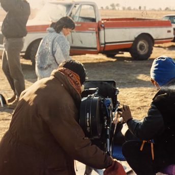 Production still of Rea Tajiri with her back to the camera. She is on set and looking into a 35mm cinema camera. Zack Winestine, the film's DOP, sits to her right. In front of them, Heather Yoshimura, an actor, runs next to two production crew. An out of focus orange truck and brown car are in the background.
