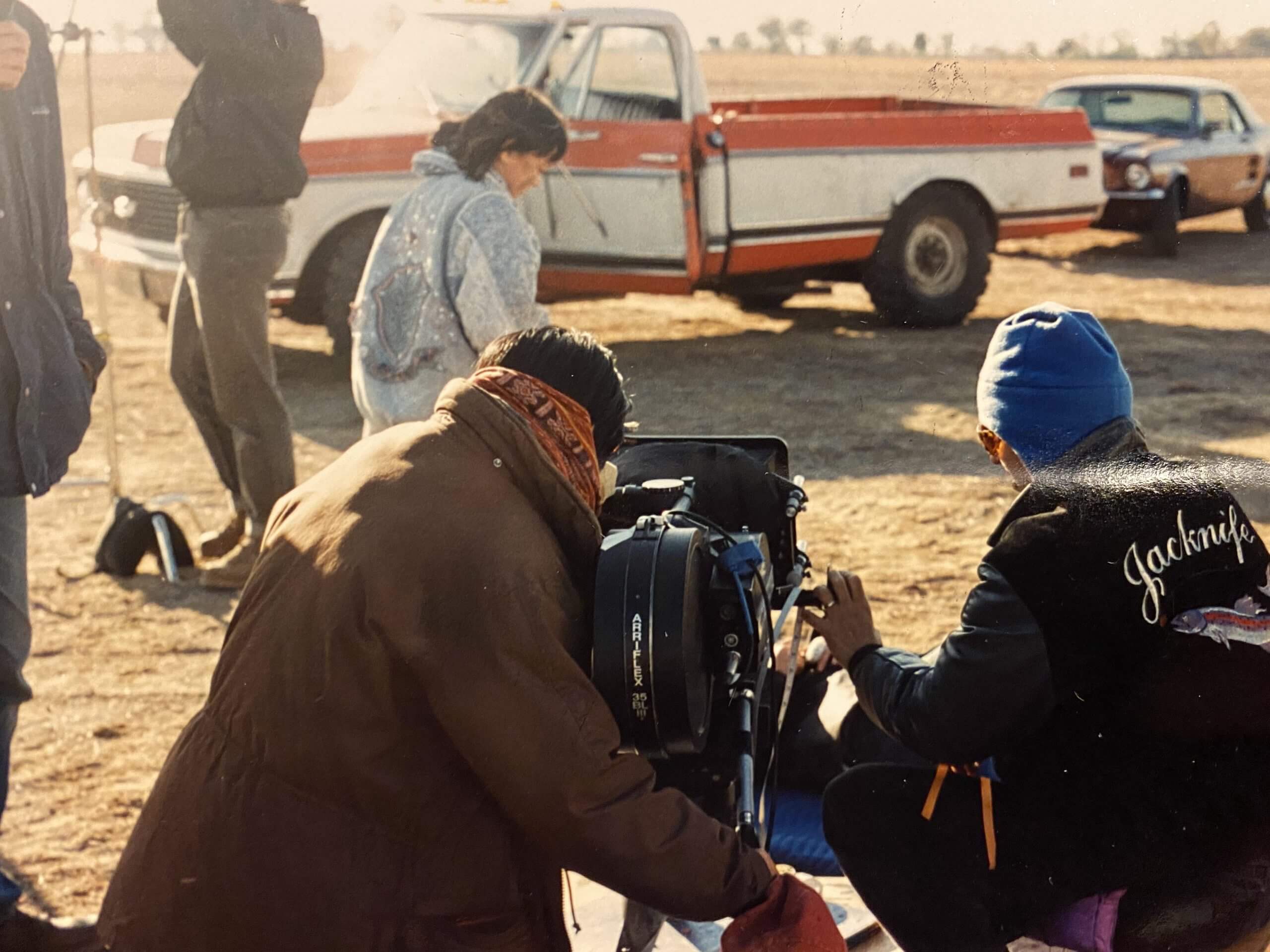 Production still of Rea Tajiri with her back to the camera. She is on set and looking into a 35mm cinema camera. Zack Winestine, the film's DOP, sits to her right. In front of them, Heather Yoshimura, an actor, runs next to two production crew. An out of focus orange truck and brown car are in the background.
