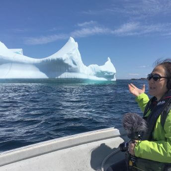 Production still of Rea Tajiri sitting in a side profile to the camera. She is wearing a bright green coat and is in a boat on the ocean. She is in front of an iceberg, wearing headphones and sunglasses. She is holding a camera and microphone with a fluffy cover.