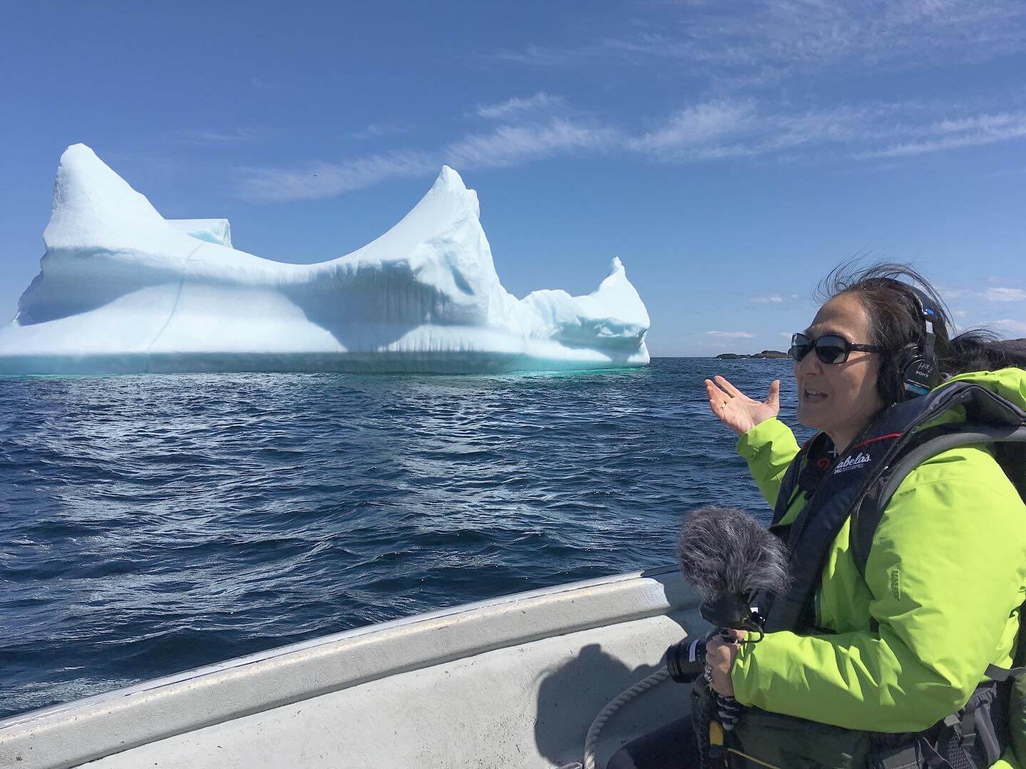 Production still of Rea Tajiri sitting in a side profile to the camera. She is wearing a bright green coat and is in a boat on the ocean. She is in front of an iceberg, wearing headphones and sunglasses. She is holding a camera and microphone with a fluffy cover.