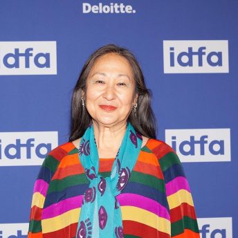 Rea Tajiri looking directly into the camera and smiling. She is standing in in front of the IDFA FILM FESTIVAL wallpaper. She is wearing a multicolored and striped shirt, with a teal scarf.