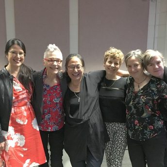 A group of women film directors stand together in a horizontal row directly facing the camera and are all smiling. From furthest left to right: Kimi Takesue is in a red dress; Barbara Hammer is wearing a floral blouse and red glasses; Rea Tajiri is in the center, wearing a black shirt; Elisabeth Subrin is standing to her right with short cropped hair; Sarah Drury is in a floral blouse; and Chet Pancake leans into frame smiling