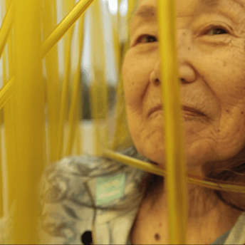 Still from Wisdom Gone Wild. An elderly Asian woman is sitting in a wheelchair, smiling, and looking into the camera. In front of her are yellow streamers.