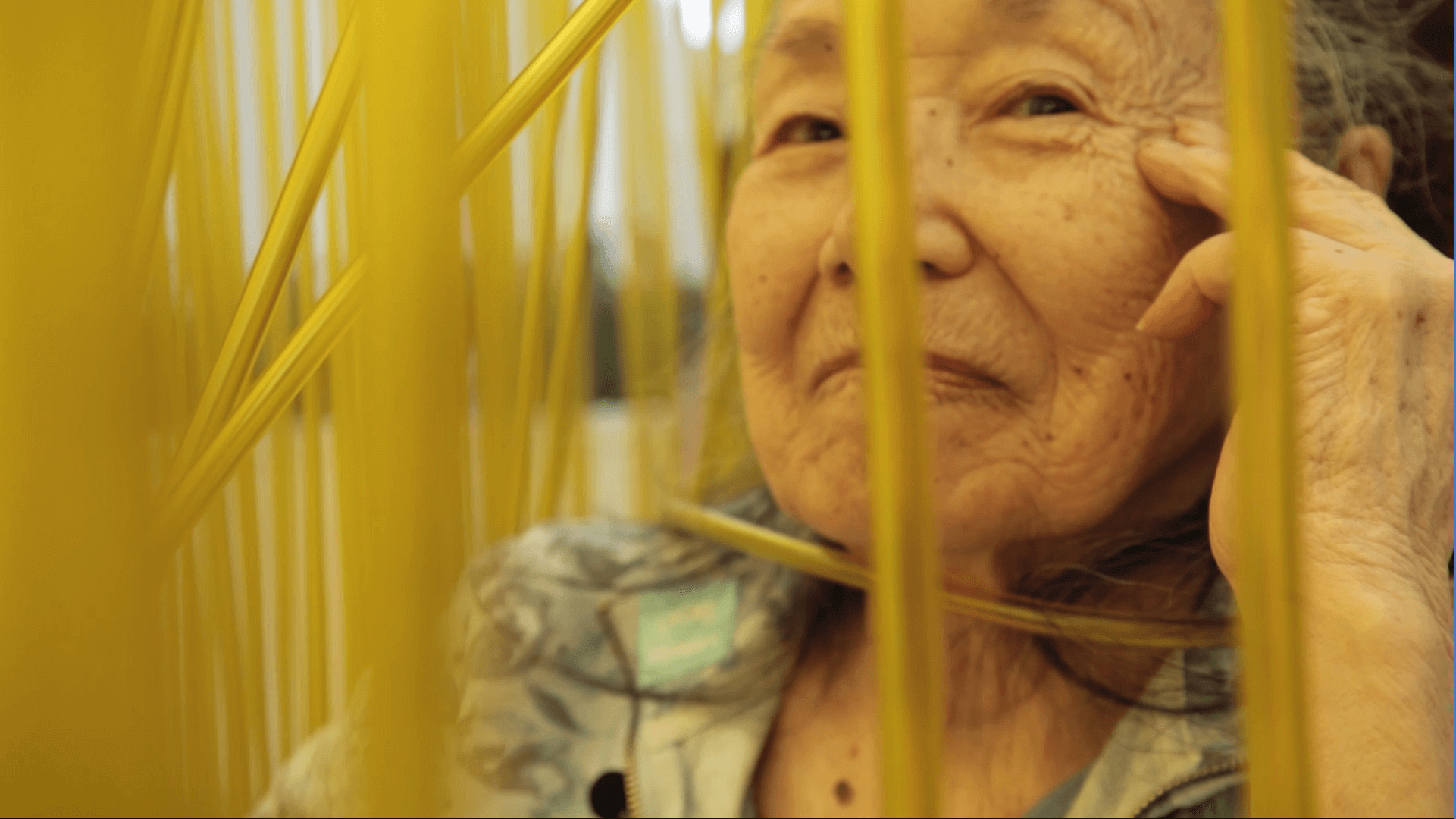 Still from Wisdom Gone Wild. An elderly Asian woman is sitting in a wheelchair, smiling, and looking into the camera. In front of her are yellow streamers.