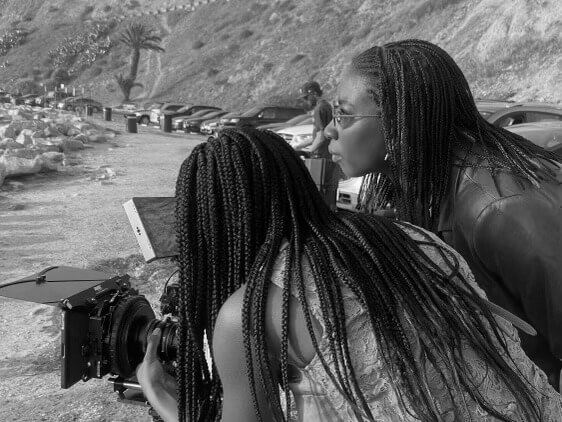 Production still of Sabaah Folayan standing in profile to the camera and wearing sunglasses. They are next to another woman with braids and a sleeveless blouse whose back is to the camera. They are both leaning forward and looking into a camera, which is pointed offscreen.