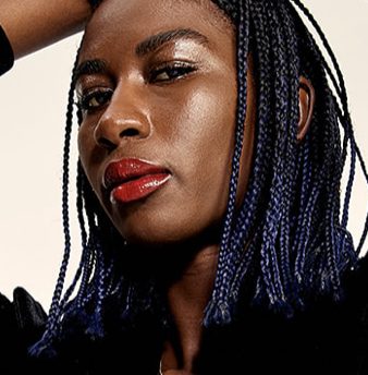 Sabaah Folayan looking directly at the camera. She is wearing blue braids and red lipstick.