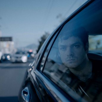 Still from Enemies of the State. A young man is looking through the side window of a driving car. Image is taken from the outside and focuses on the man, while the background is out of focus. There is a police car in the distance.