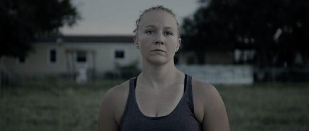 Still from United States vs. Reality Winner. A young woman, Reality Winner, is standing in a field in front of a house. She has a serious expression. The light is low and the background is out of focus.