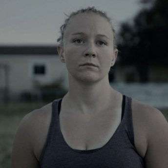 Still from United States vs. Reality Winner. A young woman, Reality Winner, is standing in a field in front of a house. She has a serious expression. The light is low and the background is out of focus.