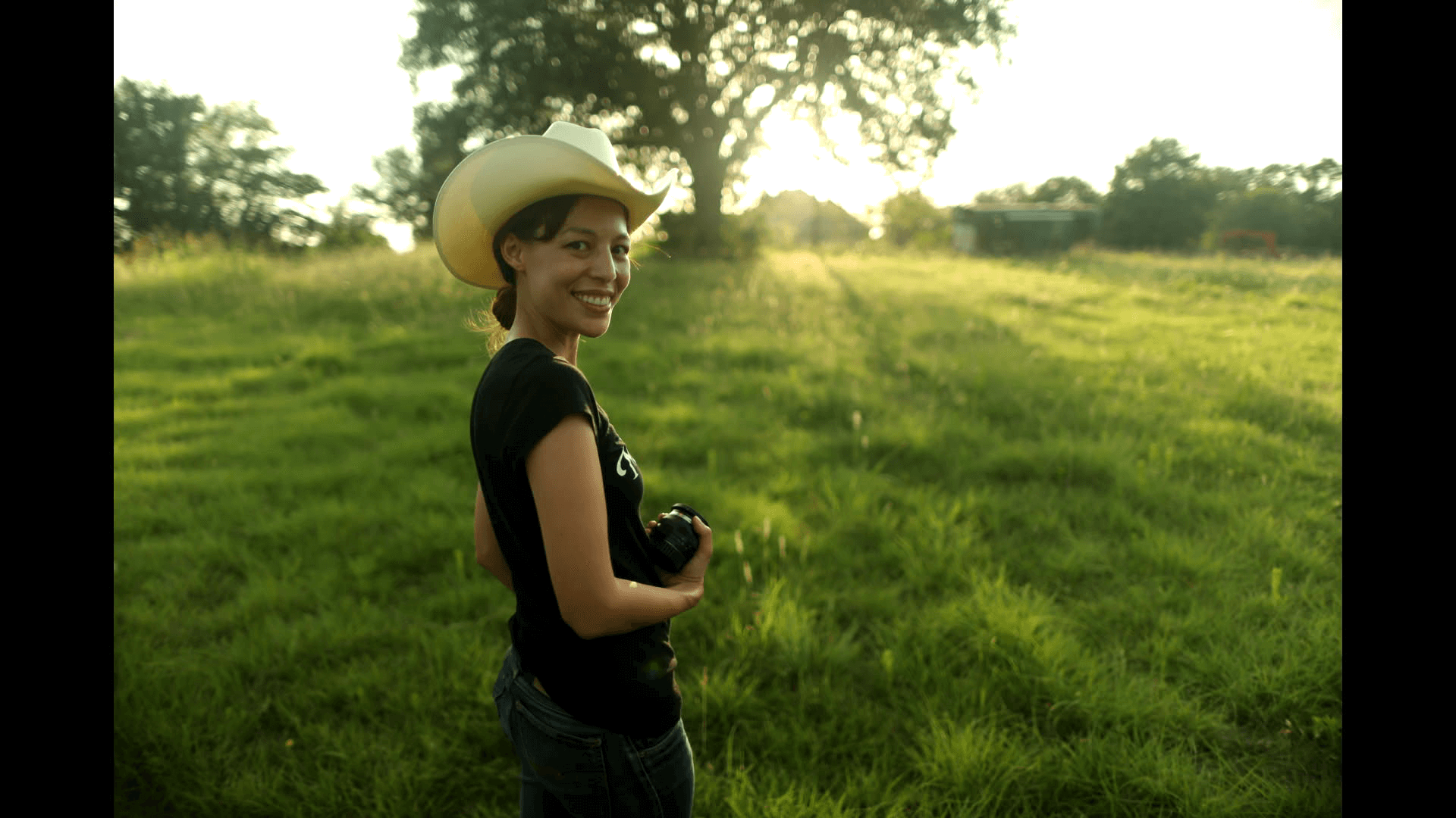 Sonia Kennebeck looking directly at the camera. She is standing in a green field with a tree in the background and is holding a photo lens.