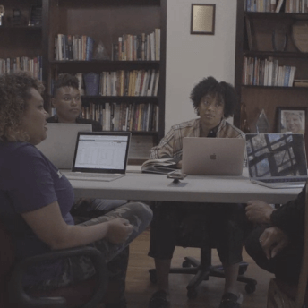 Production still of After Roe. SisterSong staff sitting around a table with their laptops open. They are turned towards one another and away from camera. There are filled bookshelves in the background with books and trophies.