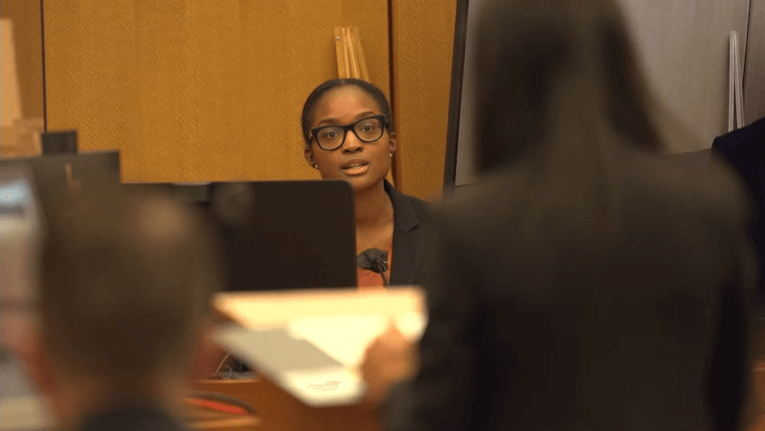 Production still of After Roe. Dr. Whitney Rice sitting and testifying in court during the SisterSong v Georgia trial. She is wearing glasses and is speaking towards a blurred woman in the foreground, who is at a podium and has her back to the camera.