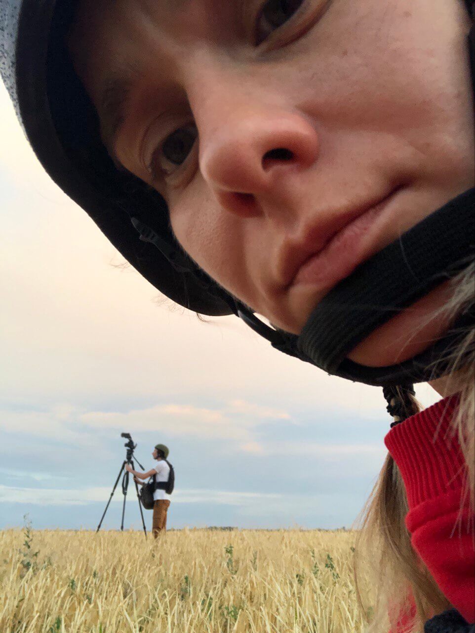 Production still of Displaced. Olha Zhurba, director of Displaced, is taking a photo of herself and is shown in a close-up. In the background is Volodymyr Usik, Director of Photography, who stands in profile and looks up at a camera. They are close to the frontline, and are both wearing flat jackets and helmets.