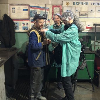 Production still of Displaced. Three men are shown in a bread factory in Mykolayiv. One man, the sound director, is mounting a mic on one of the film's subjects, Vitaly, a bread factory worker. On the wall behind them are various posters.