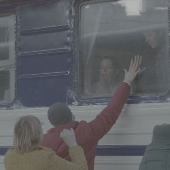 Still from Displaced. A man is saying goodbye to his wife and son through the window of the train. His hand reaches to the window from the train station platform as his family looks down.