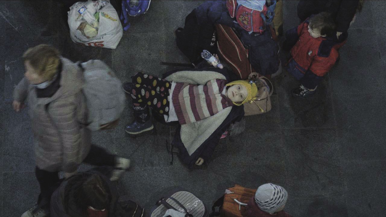 Still from Displaced. A girl is lying on family belongings at the train station. She looks directly up at the camera as other citizens walk past her.