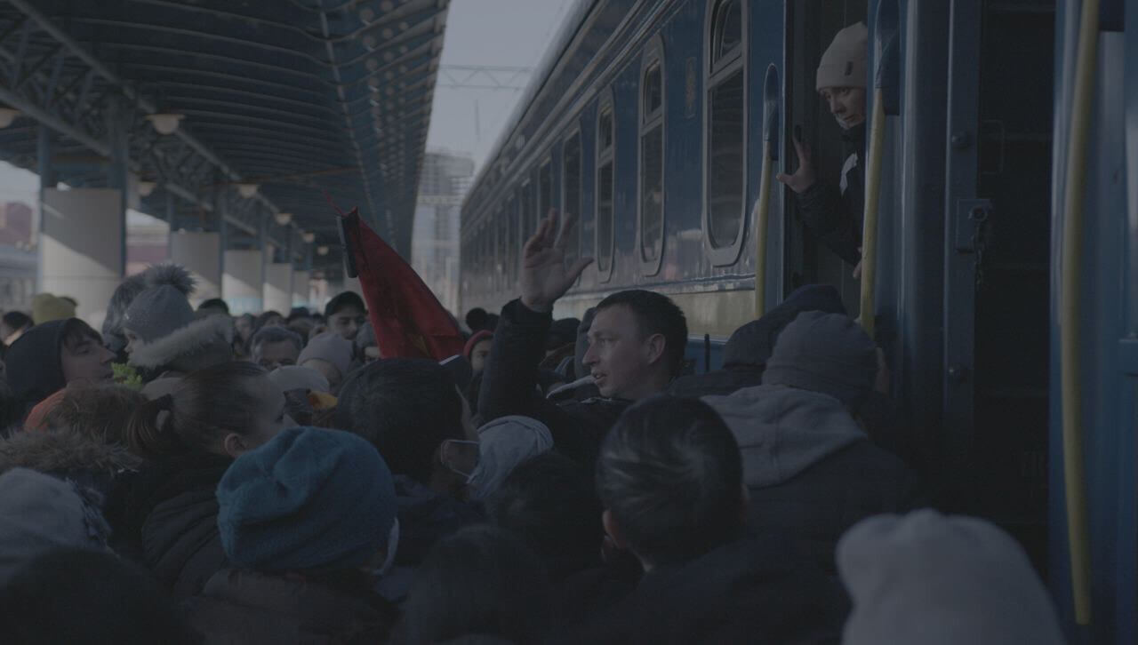 Still from Displaced. A main railway station in Kyiv, Ukraine during the first days of war. A crowd of people, who are attempting to get on an overcrowded train and evacuate the city, fills the photo. A man in profile to the camera extending his arm can be seen in the middle.