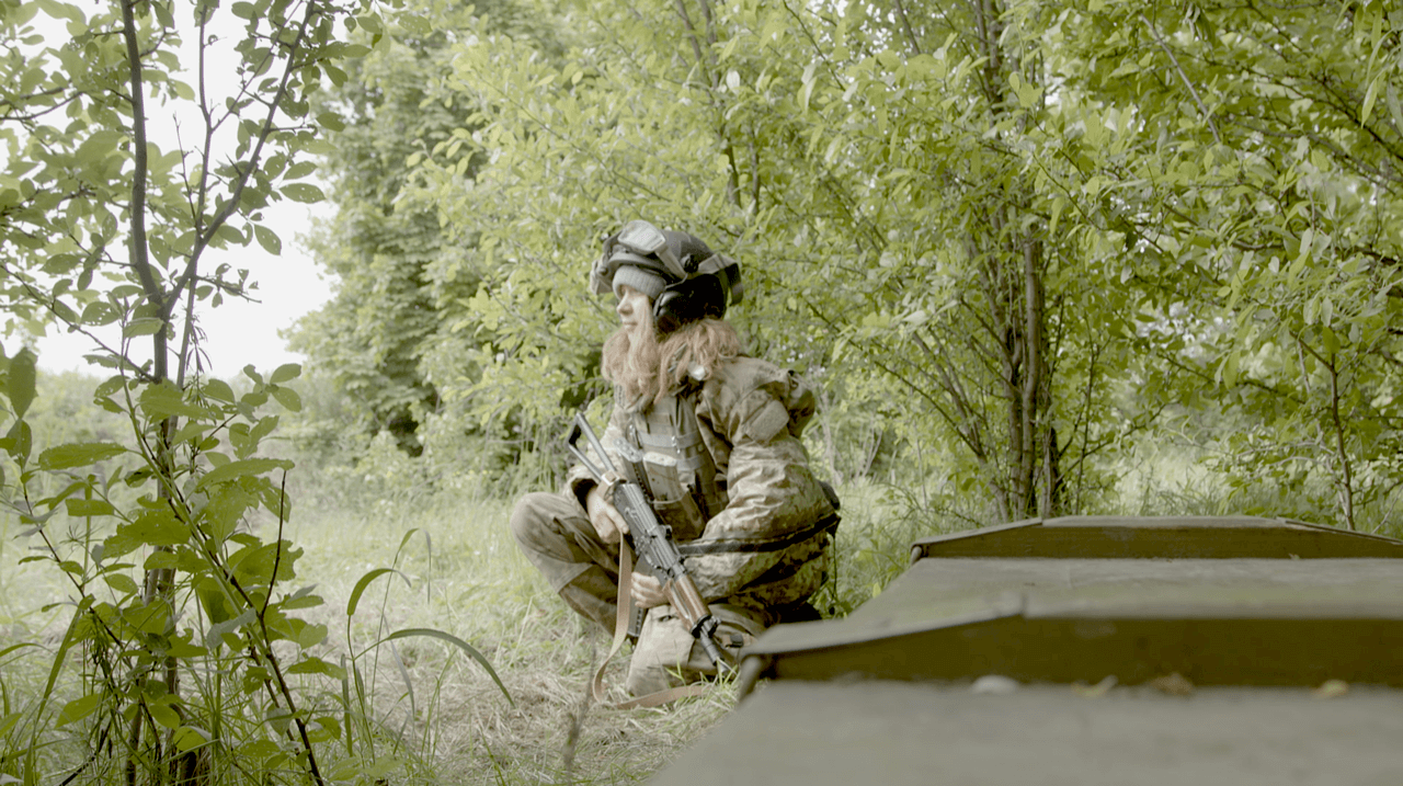 Production still of Frontline. Alisa Kovalenko, director, is squatting in a side profile to the camera. She is wearing a full military gear and holding a rifle. Various green trees surround her as she looks onward to the left.