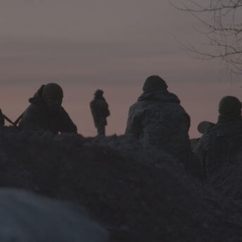 Still from Frontline. A group of soldiers is sitting on the ground, waiting, and looking on the horizon. The camera is behind them. There are mounds of dirt in the still as the camera is placed at a low angle in a trench.