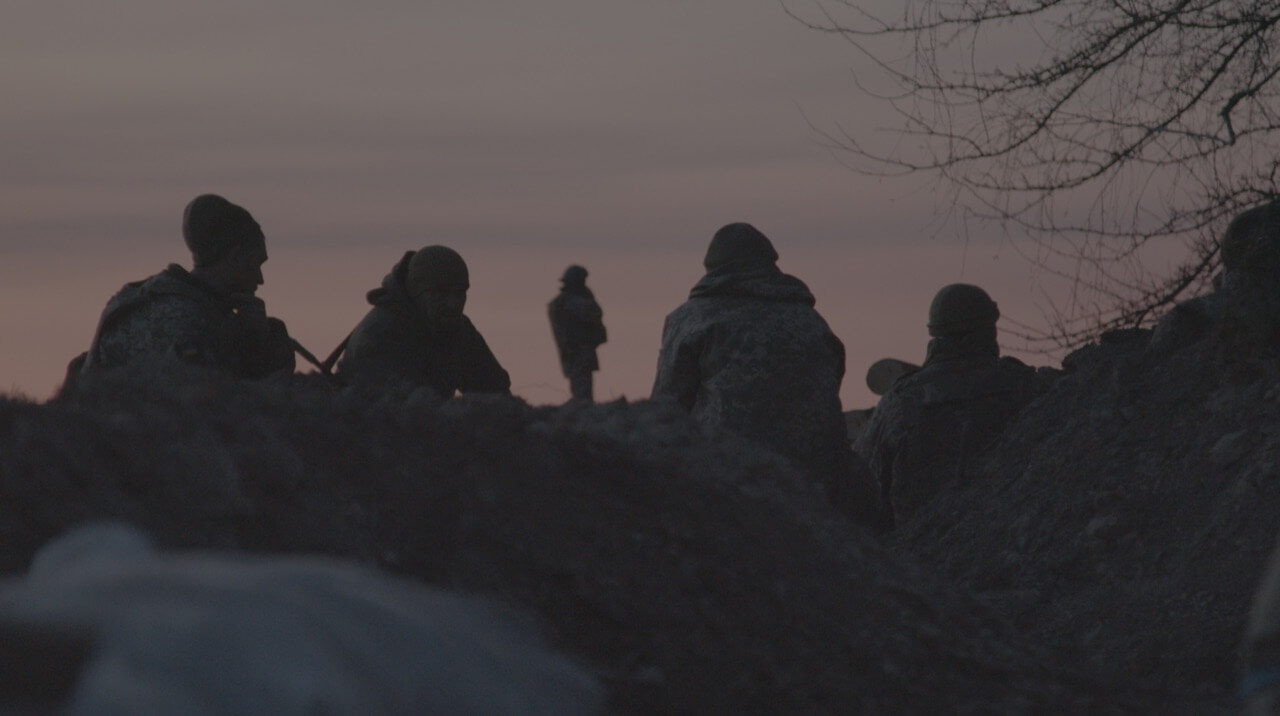 Still from Frontline. A group of soldiers is sitting on the ground, waiting, and looking on the horizon. The camera is behind them. There are mounds of dirt in the still as the camera is placed at a low angle in a trench.